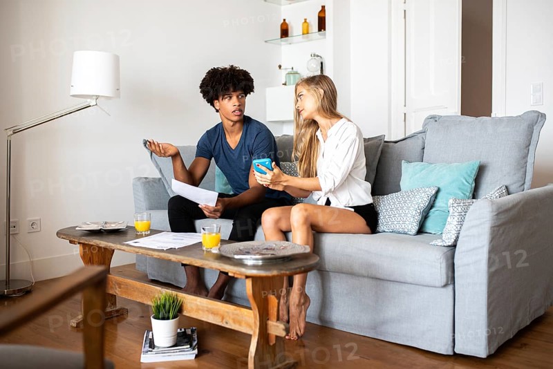 A couple is seated on a cozy gray couch, engaging in a lively discussion while holding papers and a phone. The bright living room creates a warm, inviting atmosphere