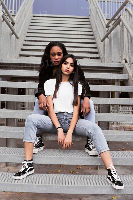 Two young women in casual outfits and sneakers sit on metal stairs. They exude confidence and style, framed by an urban backdrop, showcasing a relaxed vibe