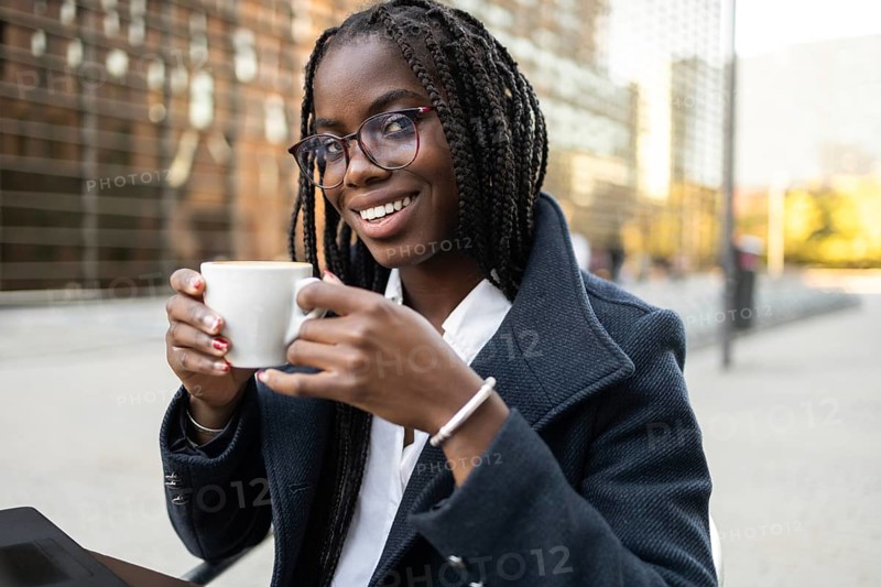 An African American businesswoman with braids, wearing glasses and a smart coat, enjoys a coffee break while working remotely in an urban setting