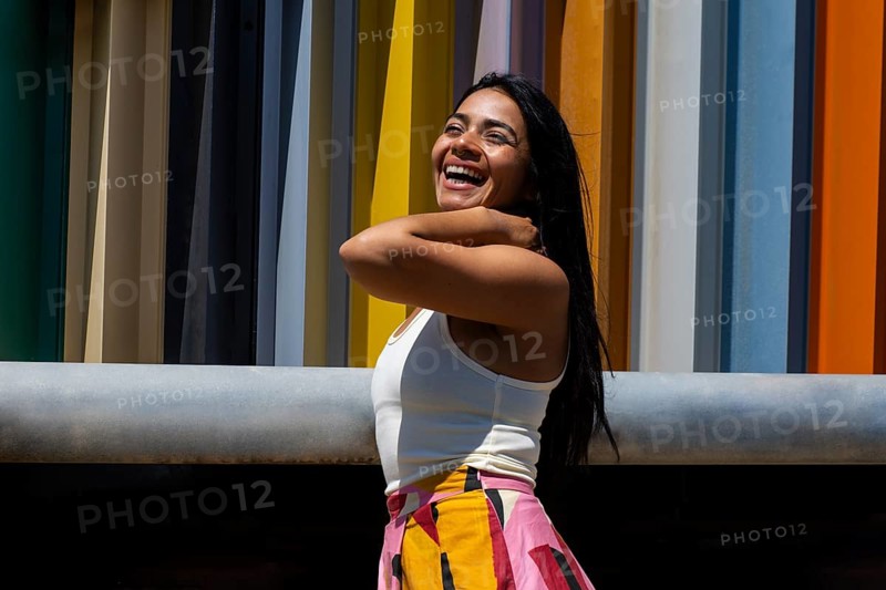A joyful woman in a vibrant outfit stands against a backdrop of modern architecture, creating a striking urban contrast. Her expression radiates happiness and confidence