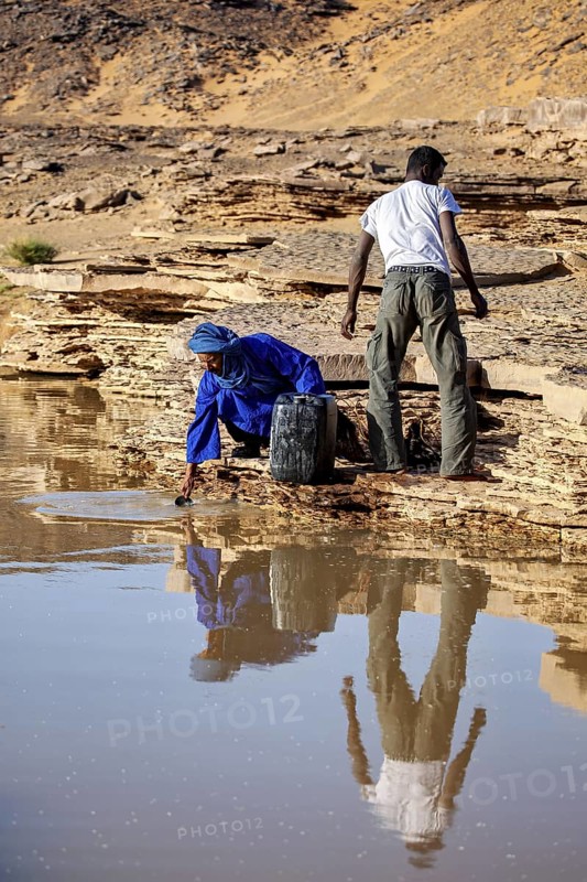 Two people drawing water from a spring in a barren desert landscape, Tuareg fetching water in the Sahara in Algeria
