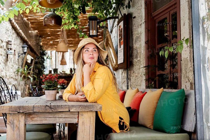 A young woman in a vibrant yellow shirt sits at a rustic cafe table, casually wearing a hat. She looks away thoughtfully, surrounded by greenery and cozy cushions