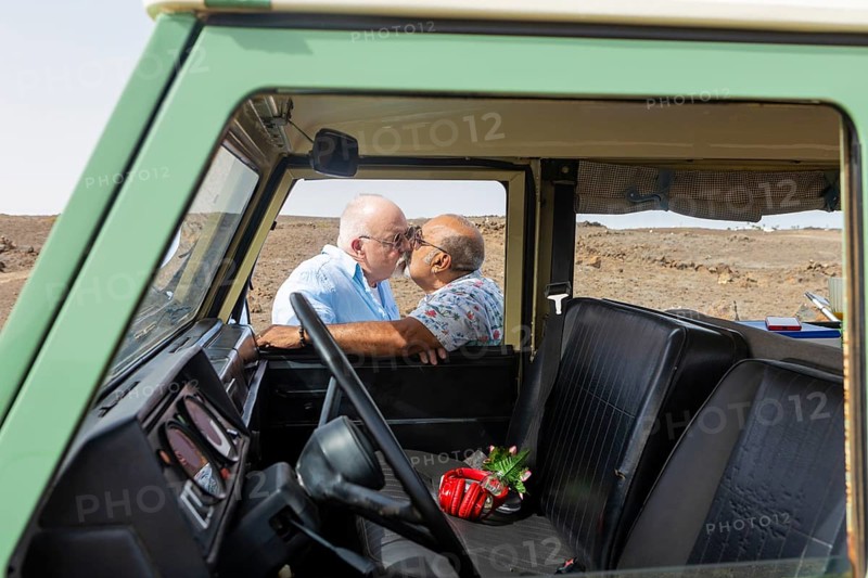 An affectionate elderly gay couple kisses inside an off-road vehicle during a camping trip, illustrating a moment of love and companionship in a serene landscape