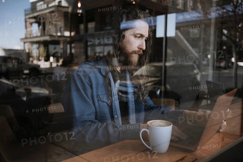 A man with long hair and a beard focuses on his laptop in a cozy coffee shop. A cup of coffee sits beside him, with reflections of the street visible on the glass