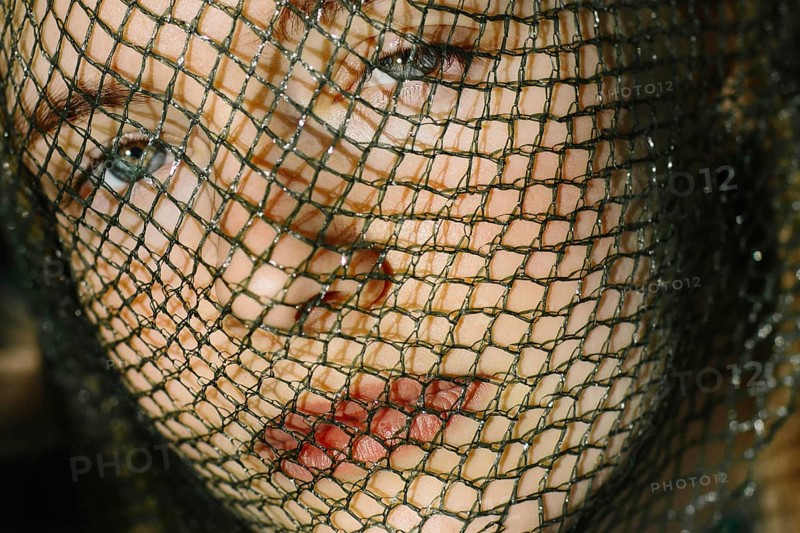 A close-up portrait of a teenager behind a textured mesh screen, capturing a sense of mystery and introspection. The soft lighting highlights their youthful features and contemplative expression