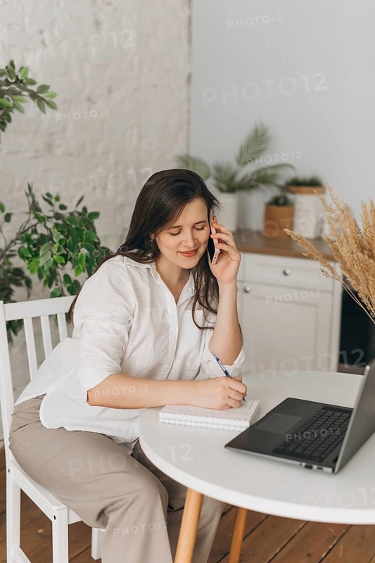 A nutritionist in a home office setting consults with a client over the phone. She takes notes on a notepad beside a laptop, surrounded by indoor plants, creating a serene workspace