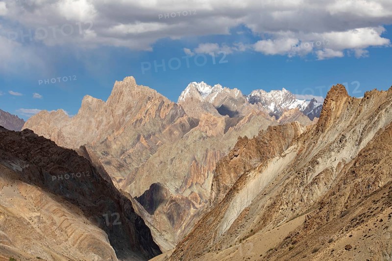 Sunlit peaks of the Zanskar Valley, Ladakh, India, display geological splendor with sharp, rugged formations. The dramatic landscape is framed by a vibrant blue sky and wispy clouds