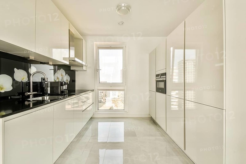 A bright modern kitchen featuring sleek white cabinets, glossy countertops, and a window with a city view. The design emphasizes minimalism and functionality