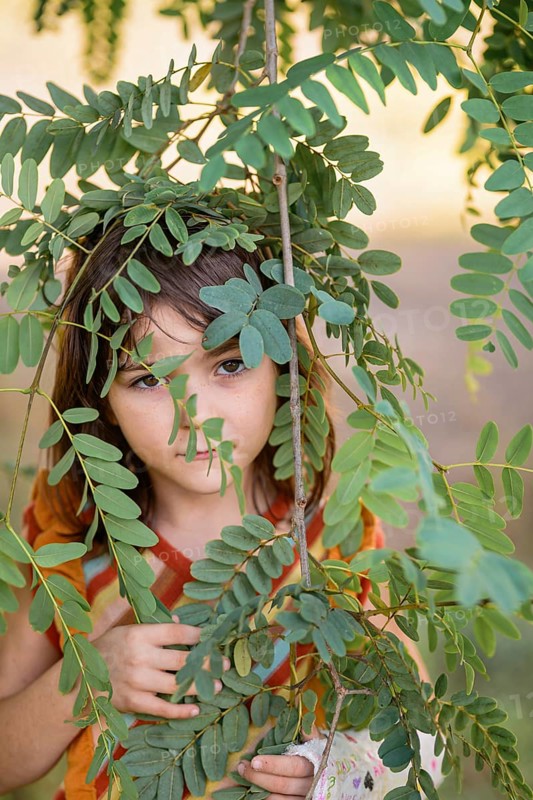 A young girl peeks through vibrant green leaves, offering a glimpse of curiosity and connection with nature. The scene captures a moment of playful exploration