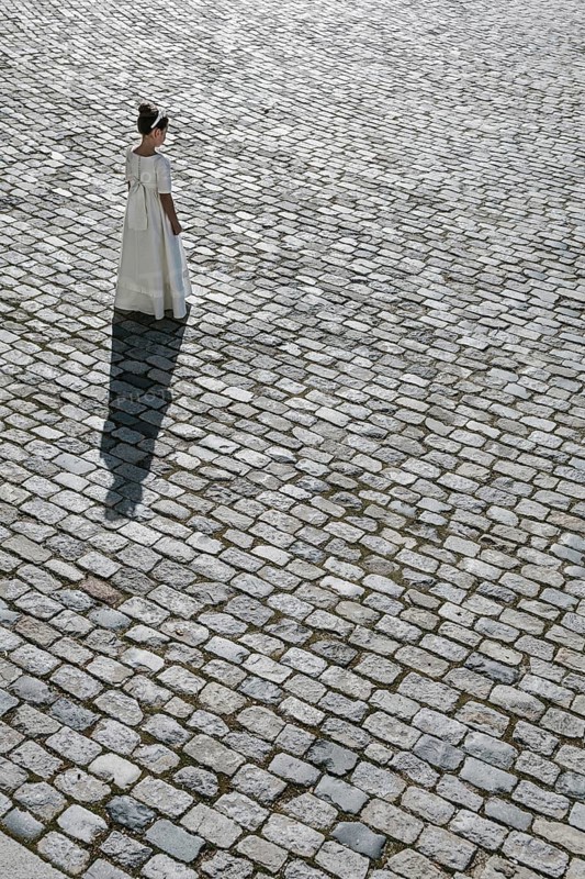 A young girl stands alone in a white First Communion dress, casting a long shadow on a cobblestone path. The scene evokes a sense of innocence and tradition