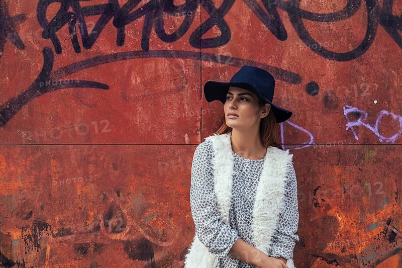 A woman wearing a fashionable black hat and casual outfit leans against a vibrant graffiti-covered wall. Her relaxed pose and thoughtful expression embody urban style