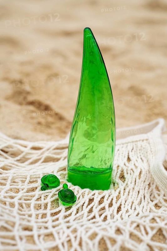 On a sandy beach, a green aloe gel tube stands upright on a white mesh fabric, accompanied by three small green capsules. Ideal for skincare routines focused on natural ingredients