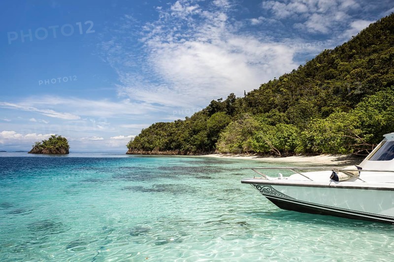 Overlooking the tranquil beaches and turquoise waters of Raja Ampat, this image showcases a boat moored near leafy, secluded islands under a bright, cloudy sky