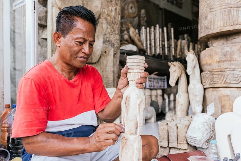 An Indonesian man skillfully carves a detailed sculpture in a studio filled with wooden art pieces, showcasing traditional craftsmanship and dedication to art