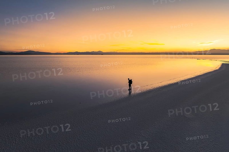 A lone traveler stands at the edge of a vast salt flat in Salinas Grandes, Salta, Argentina, as the sky is painted with the warm hues of a serene sunset reflecting on the water