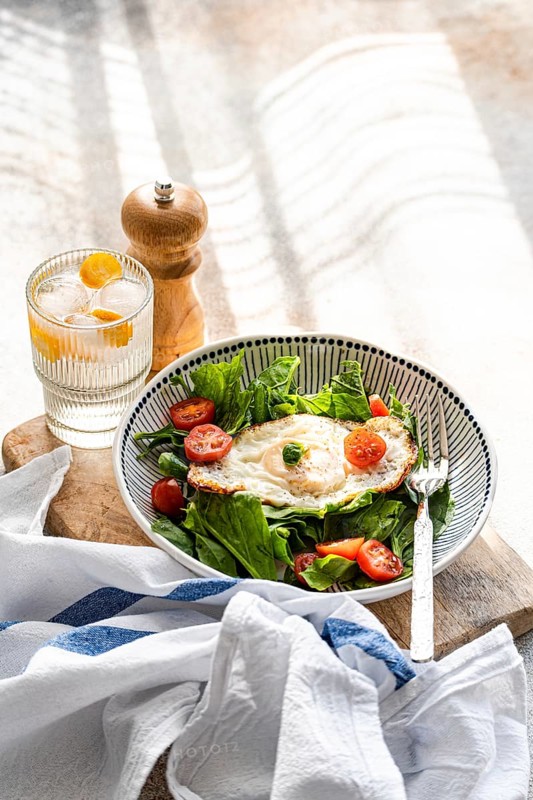 Top view of a refreshing healthy lunch featuring a plate of fresh spinach leaves topped with cherry tomatoes and a fried egg, served alongside a glass of mineral water with ice and kumquat slices