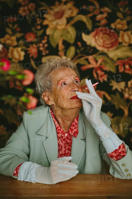 An elderly woman in a vintage setting, wearing elegant attire, looks thoughtfully with a pink candle. The floral background adds to the nostalgic and classic vibe