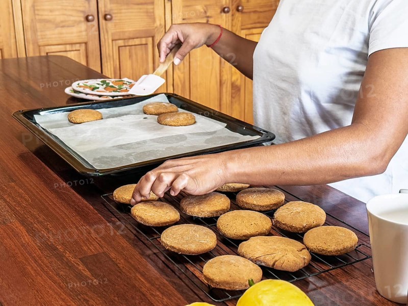 A woman bakes fresh cookies on a wooden table. The scene shows cookies placed on a cooling rack beside a baking tray, with ingredients and tools nearby, capturing the essence of home baking