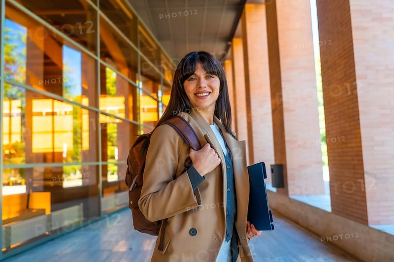Young woman carrying a backpack and tablet, confidently smiling while walking on a modern university campus, representing education, career, and academic success