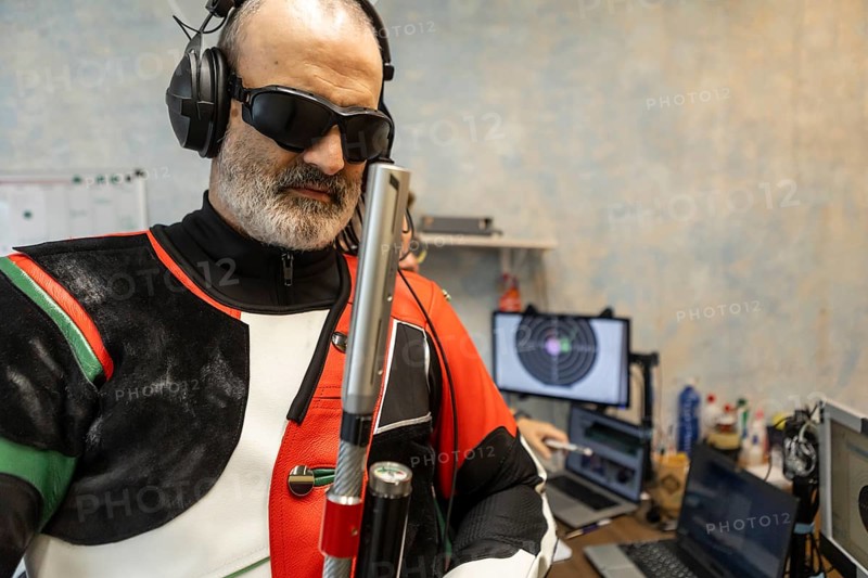 A blind Olympic shooter in protective gear stands ready, showcasing determination and skill. The background features target screens and equipment, highlighting the sport's precision