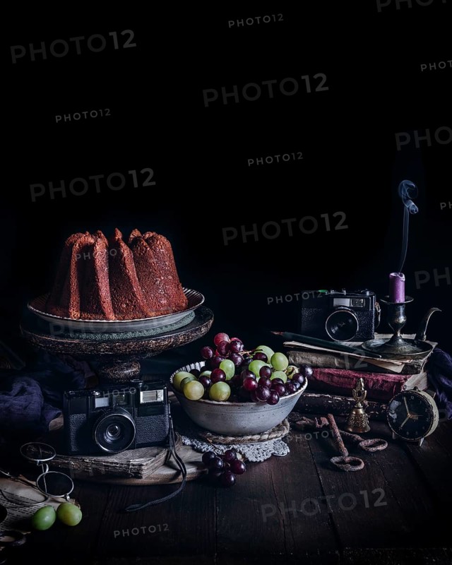 A captivating vintage still life featuring a cake on a stand, surrounded by antique cameras, a bowl of grapes, old books, keys, and a candle, creating a nostalgic ambiance
