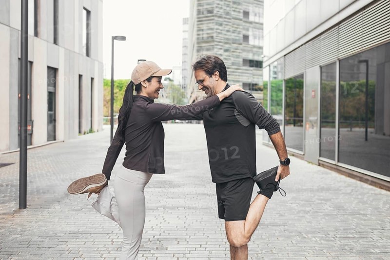 A couple dressed in sportswear performs a leg stretch on a city street, supporting each other with smiles. The backdrop features modern architecture and greenery