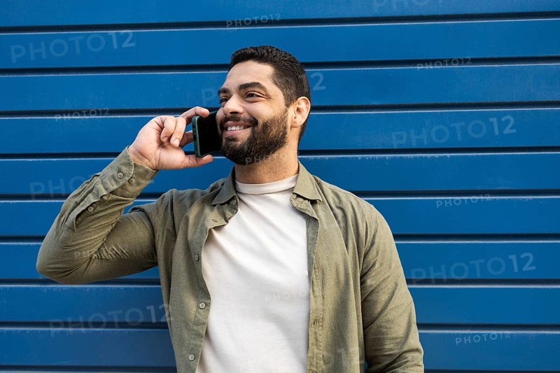 A cheerful man speaks on his smartphone against a modern blue wall backdrop. He wears a casual green shirt and a beige t shirt, exuding a positive and relaxed vibe