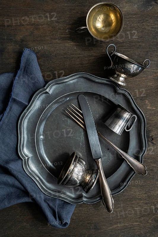 A rustic arrangement of vintage metal tableware, featuring a plate, fork, knife, and two small cups on a dark wooden surface, complemented by a blue napkin