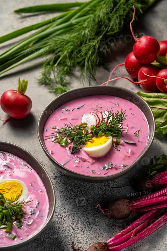 A vibrant bowl of cold beetroot soup topped with chopped herbs, a slice of boiled egg, and fresh radishes. The bright pink soup is complemented by fresh greens