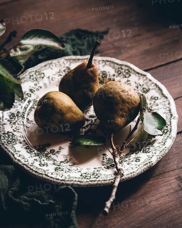 Three ripe pears rest on an intricately patterned plate, placed on a rustic wooden table. Natural lighting enhances the warm, earthy tones, creating an inviting atmosphere