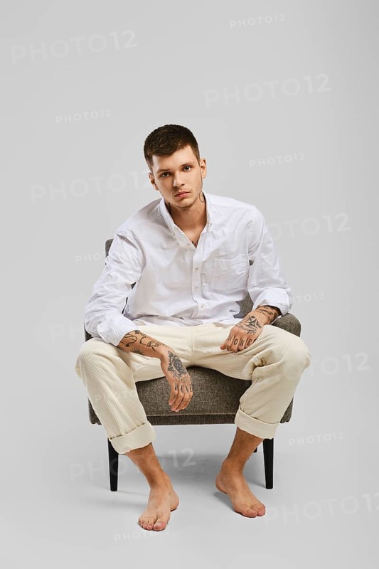 Full length studio portrait of young barefoot man in white shirt and ivory trousers sitting in armchair