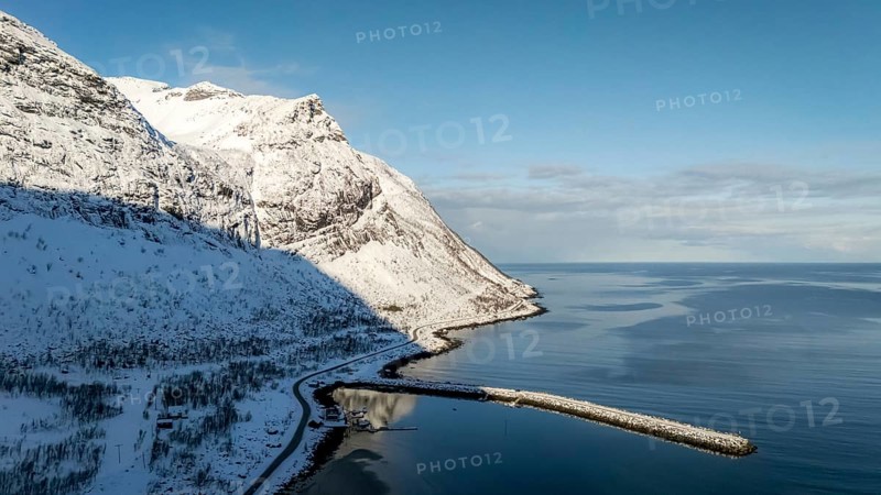 A stunning aerial view captures the serene Nordic coastline, featuring majestic snow-covered mountains meeting the deep blue sea. Clear skies add to the breathtaking scenery, Skaland, Troms, Norway