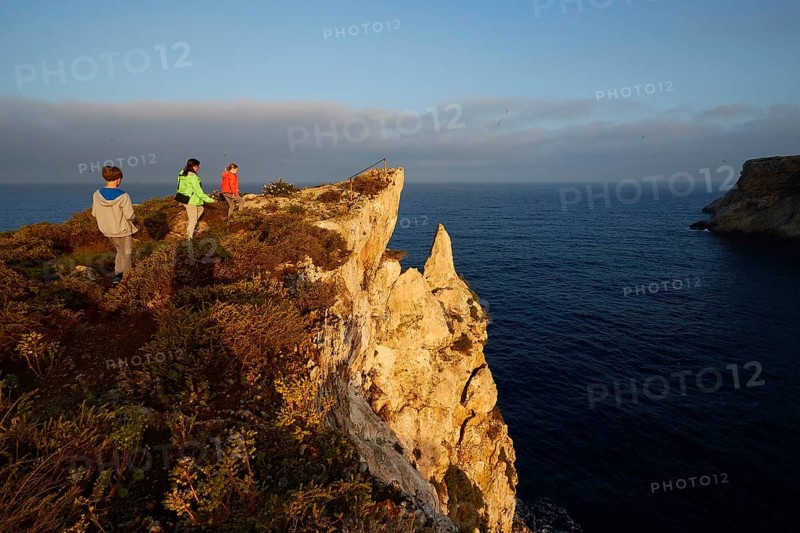 Three people explore a cliff overlooking the sea at sunrise, morning light, Tsichli Babas islet, Navarino Bay, Pylos, Peloponnese, Greece