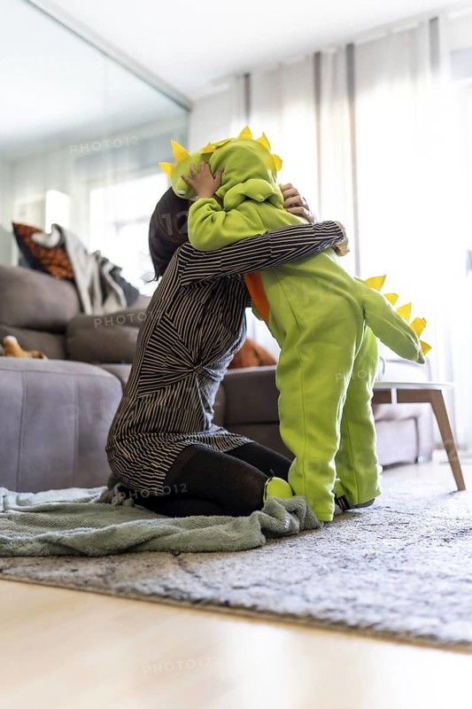 Mother kneeling on carpet and tenderly hugging her son wearing a dinosaur costume, in a cozy home environment