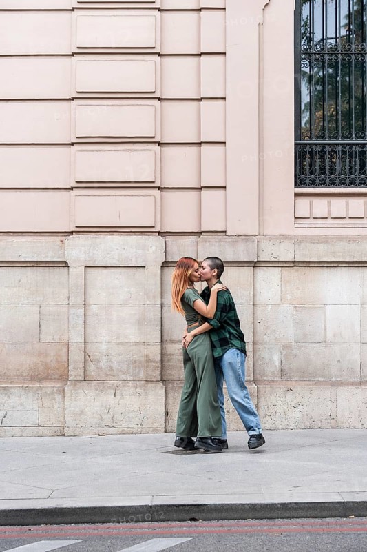 A loving lesbian couple shares a tender kiss on a city sidewalk, framed by elegant historic architecture. Their affectionate embrace captures a moment of love, connection, and timeless romance