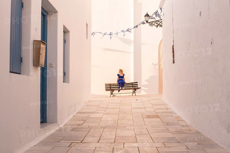 A woman in a blue dress sits on a bench in a sunlit alley of a Greek village in Plaka, Milos island. White walls and blue accents create a serene atmosphere, while flags add a festive touch