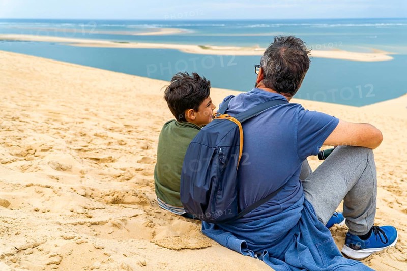 A father and son sit on the dune of Pilat, overlooking the serene ocean. The moment captures tranquility and bonding against a backdrop of blue waters and expansive sky