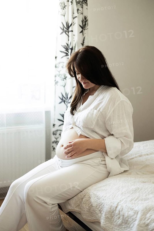 A pregnant woman gently cradles her belly, reflecting on the upcoming journey into motherhood. She sits peacefully on a bed in a softly lit room, symbolizing growth and anticipation