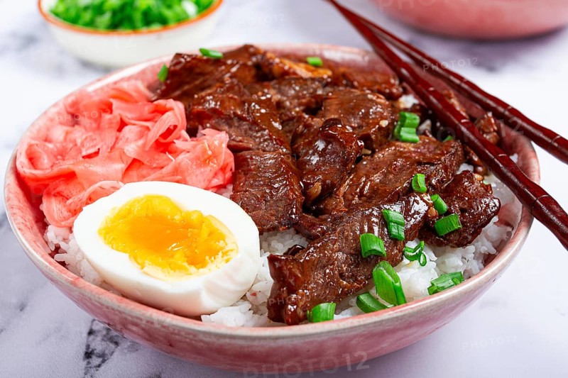 Gyudon Japanese beef with rice, pickled ginger, boiled egg slice, garnished with green onions, in a bowl with chopsticks