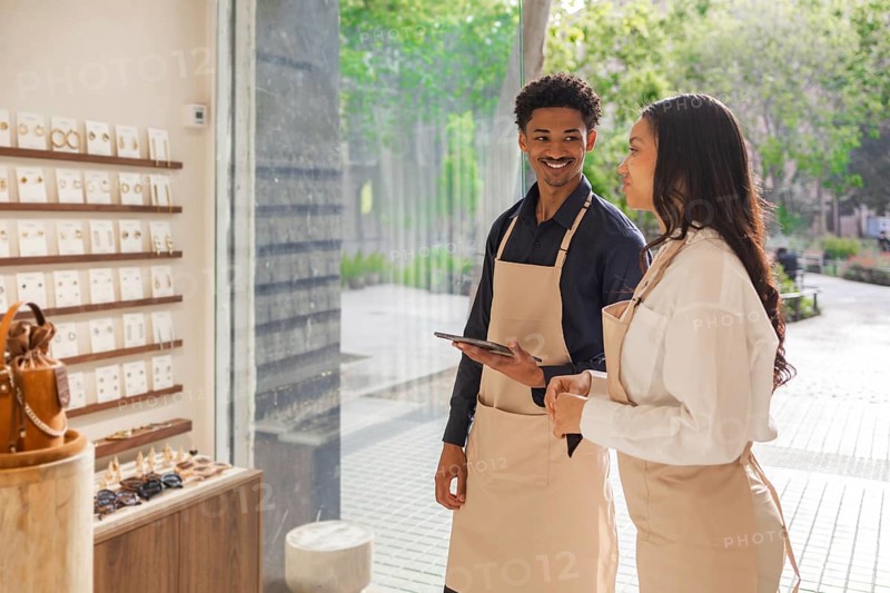 Two store employees in aprons engage in a conversation about product display in a stylish, sunlit bag store. Both are smiling, creating a welcoming atmosphere for customers