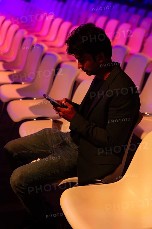 A indian man in a dark blazer and jeans sits in an empty, illuminated auditorium at a conference of AI in Amsterdam. Engrossed in their smartphone, they are surrounded by a vibrant, colorful glow