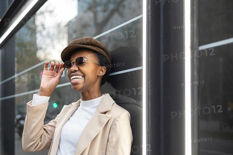 A stylish black woman wearing sunglasses and a hat enjoys a moment in the city Her fashion and smile radiate confidence, capturing an urban vibe and vibrant personality