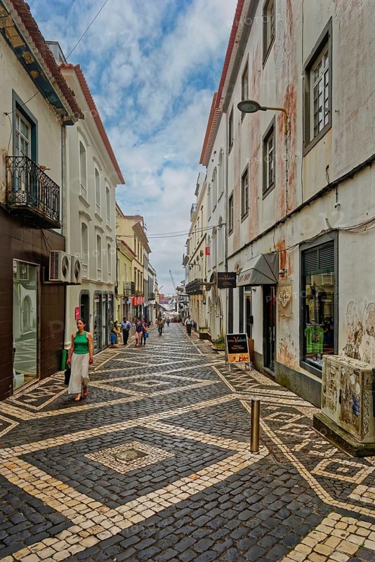 People walk along a narrow cobbled street lined with historic buildings and cafés under a clear sky, Ponta Delgada, Sao Miguel Island, Azores, Portugal
