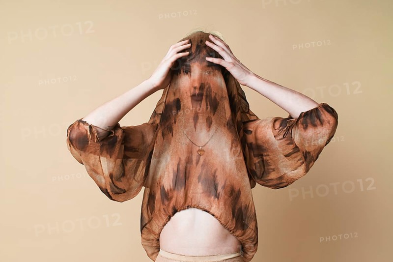 A young woman stands against a beige background in a studio, enveloped in a sheer, flowing top that she drapes over her head while looking intently at the camera