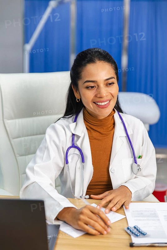 A female doctor with a stethoscope around her neck sits at a desk, writing notes and smiling. The bright office background creates a professional and welcoming atmosphere