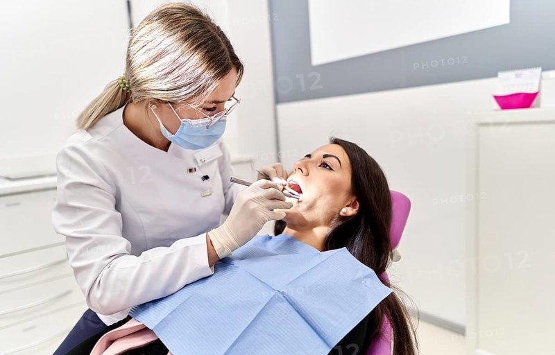 A professional dentist in a white coat and mask examines a patient's teeth at a dental clinic. The patient relaxes in a chair, showcasing a hygienic and caring environment