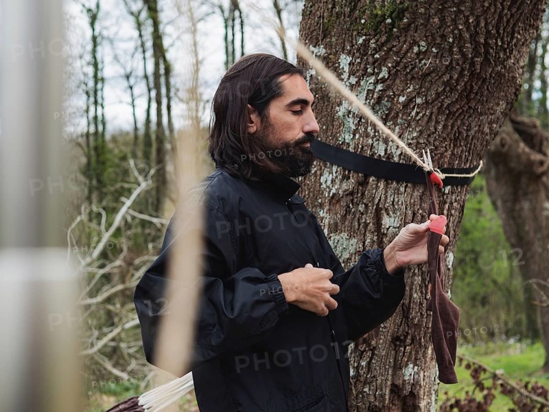 A bearded man in a black jacket hangs a piece of clothing on a tree using a rope. The peaceful outdoor setting, surrounded by trees, suggests a camping or survival scenario