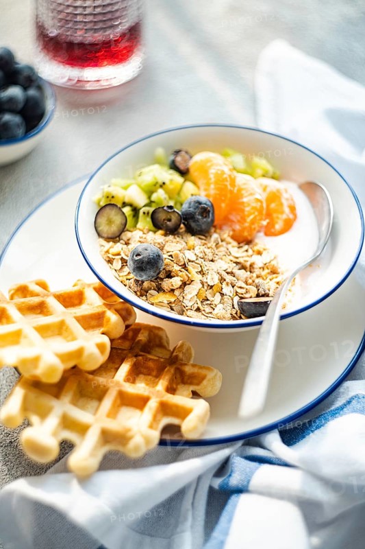 A nutritious breakfast bowl filled with granola, yogurt, blueberries, kiwi, and tangerine slices, accompanied by crispy waffles, served on a white plate with a spoon