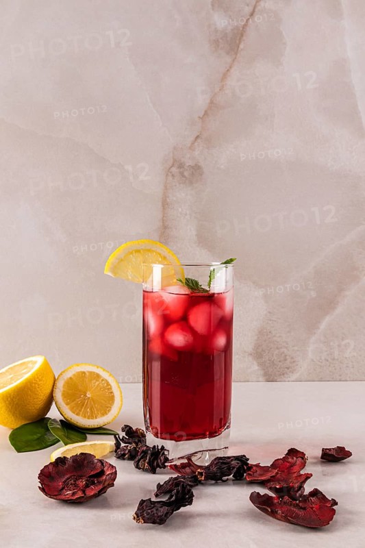 A chilled hibiscus tea in a glass with ice, garnished with a lemon slice and mint leaves. Surrounded by dried hibiscus petals and fresh lemon, set against a neutral background