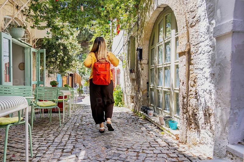 A female tourist with a red backpack strolls through a charming, sunlit cobblestone street. Lush greenery and pastel-colored chairs create a picturesque travel scene